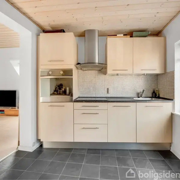 Et moderne køkken med lyst træmøblement og kogeplade under emhætte; indbygget i væg; tilstødende stue med trægulve og lav opbevaringsreol med tv. Vindue til højre.