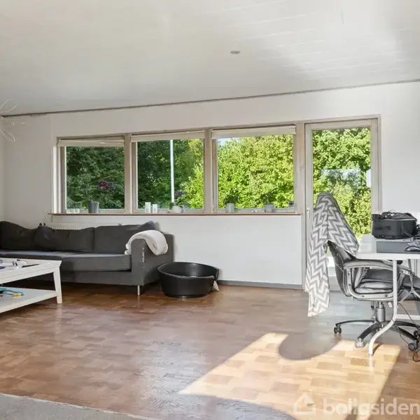 Sofa står i et hjørne ved siden af et lavt bord, der rummer en kop. Ved siden af er et skrivebord med stol. Store vinduer viser trætoppene udenfor.