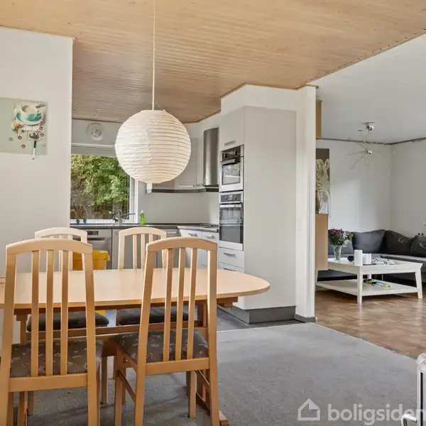 Et spisestuebord med træstole står i en åben stue-køkken-plan. En rund lampe hænger over bordet. I baggrunden ses et gråt sofaarrangement og store vinduer.