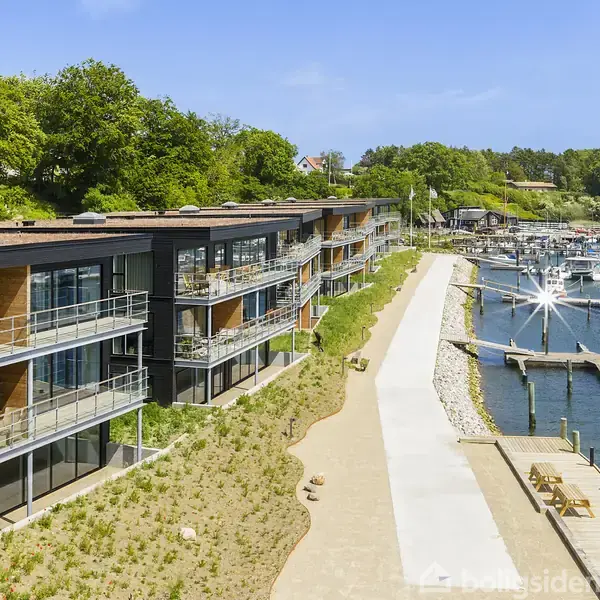 Moderne lejlighedskompleks med altaner vender mod marina, hvor lystbåde er fortøjet. Grønt område omkranser bygningen, og en gangsti snor sig ved vandet under en klar blå himmel.