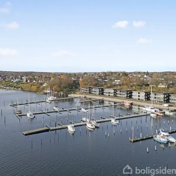 Flere både ligger fortøjet ved en marina, omgivet af en by med skovklædte områder. Moderne bygninger står langs kystlinjen, og det rolige vand strækker sig ind i horisonten under en blå himmel.