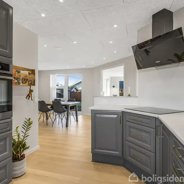 Køkken borde med grå skabe og indbygget sort ovn; rummet har lyse trægulve. I baggrunden er et spiseområde med sorte stole og et maleri på væggen.