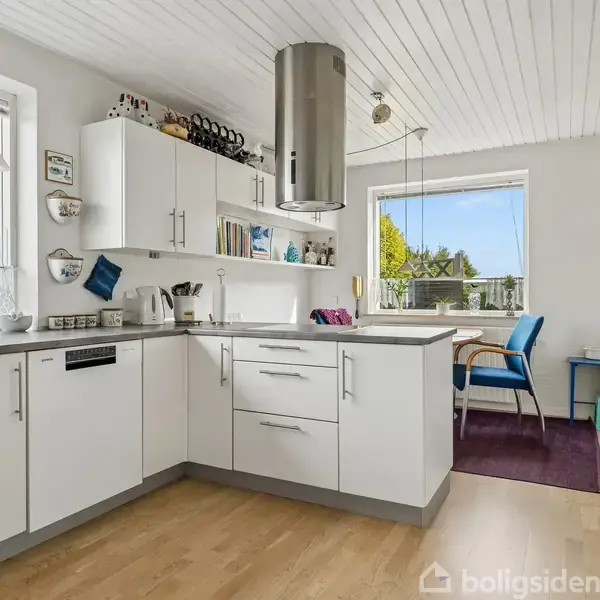 Lyst køkken med hvide skabe, grå bordplader og en stål-emhætte. To store vinduer giver udsigt til en have. Spisebord med blå stole ses i baggrunden.