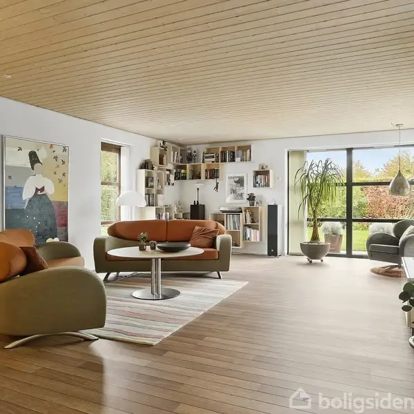 Stue med moderne møbler; to lænestole og en sofa står omkring et sofabord på et tæppe. Hylder fyldt med bøger på væggen; store vinduer giver udsigt til haven.