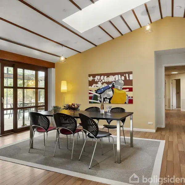 Spisebord med seks stole står på et tæppe i en stue med trægulv, højt til loftet og stor vinduesparti mod en terrasse. Vægbillede og gulvlampe bagved.