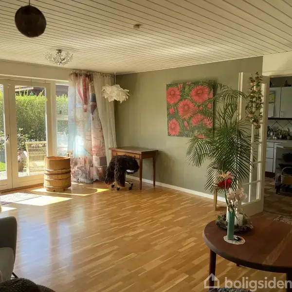 En stue med trægulv har en åben dobbelt glasdør til haven. Sollys strømmer ind. Der er et kunstværk af blomster på væggen og en plante ved siden af et bord.