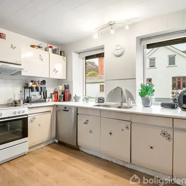 Køkken med hvide skabe dekoreret med sommerfugle og blomster. Komfur og ovn står på venstre side. To vinduer giver udsigt til nabohuse. Køkkenet er lyst og velorganiseret.