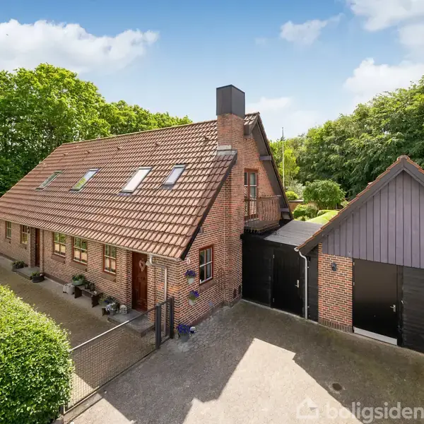 Murstenshus med rødt tegltag, mange tagvinduer og tilhørende garage. Huset ligger i en grøn have med frodige træer og indkørsel. Skyfri blå himmel over huset.
