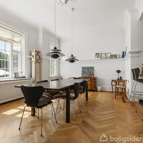Et spisebord med sorte stole står i et elegant rum med trægulv og store vinduer, der lader lys strømme ind. Rummet indeholder også en bar med høje stole.