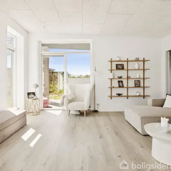 Stue indrettet med en beige sofa, rund hvidt sofabord og væghængt TV. En lænestol står nær en åbning til terrasse med udsigt til grøn natur. Væghylde med dekorative genstande.