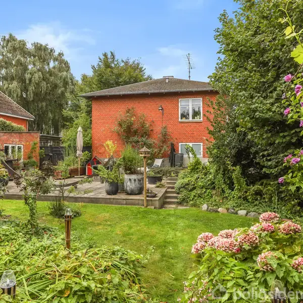 Rødt murstenshus omgivet af frodig have, med grønne planter og blomster. En terrasse med krukker og havemøbler ses foran huset. Klar blå himmel i baggrunden.
