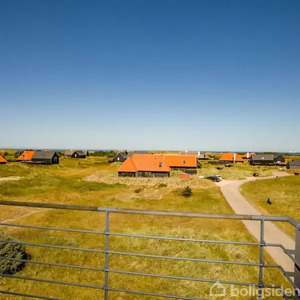 Tagterrasse udsigt viser huse med røde tage spredt over en grøn natur i et sommerhusområde. En snoet betonsti fører forbi, mens himlen er klar og blå.