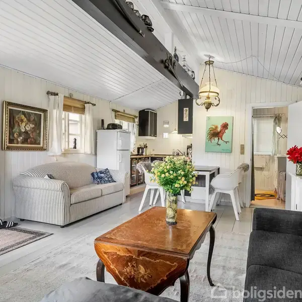 Stue med sofagruppe, træbord og blomster i vase. Lyst træinteriør og kunst på væggen. I baggrunden er der et åbent køkkenområde med spiseplads.