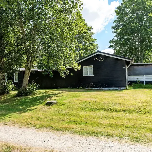 Et mørkt træhus står stille omringet af en grøn have med træer og græsplæne. En grussti snor sig i forgrunden under en skyet himmel.