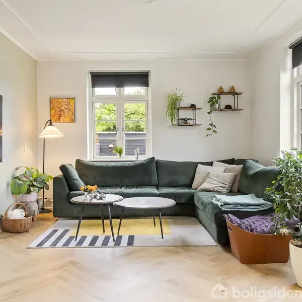 Grøn sofa står i en stue ved to vinduer, omgivet af planter og dekorationsgenstande på hylder. Et tv hænger på væggen til venstre, og der er et tæppe på gulvet.