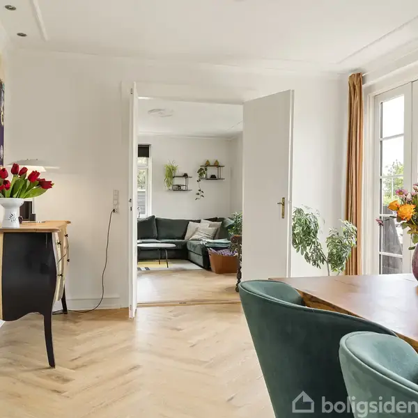 Stue med spisebord omgivet af grønne stole, vase med blomster, åbne døre til stue med sofa; lyse vægge, store vinduer, og trægulv. Tulipaner står på et sort møbel.