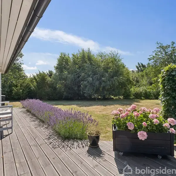 Træterrasse med hvidmalede møbler langs husets væg. Lavendel og blomsterbed pryder kanten. Haven er grøn med buske og træer under en klar blå himmel.