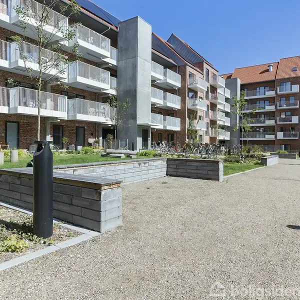 Boligblokke med altaner står omkring en grussti i en velplejet have. Der er bænke, planter og en skraldespand. Klar blå himmel skaber en solrig atmosfære.