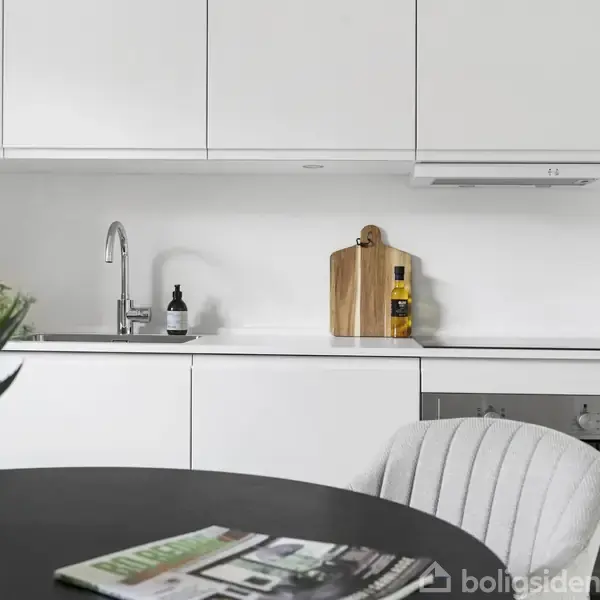 Et moderne køkken med hvide skabe, en vask med sæbedispenser og skærebræt ved siden af en indbygget ovn. En sort bordplade med en plante og et magasin i forgrunden.