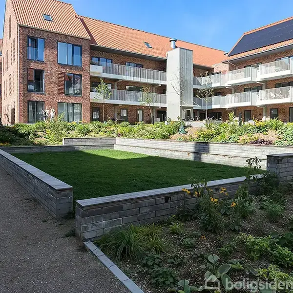 Et moderne lejlighedskompleks omkranser en grøn indergård med velholdt græsplæne og blomsterbede. Bygningerne er bygget af røde mursten og har altaner med glasrækværk.