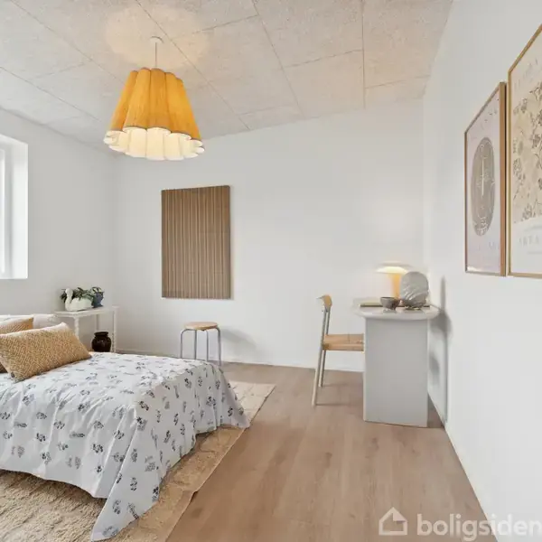 Et lyst soveværelse med en dobbeltseng dækket af blomstret sengetøj. En skrivebord med en lampe og stol står mod den hvide væg. En stor orange loftlampe hænger i midten, og vinduet til venstre slipper naturligt lys ind. På væggen hænger to plakater.