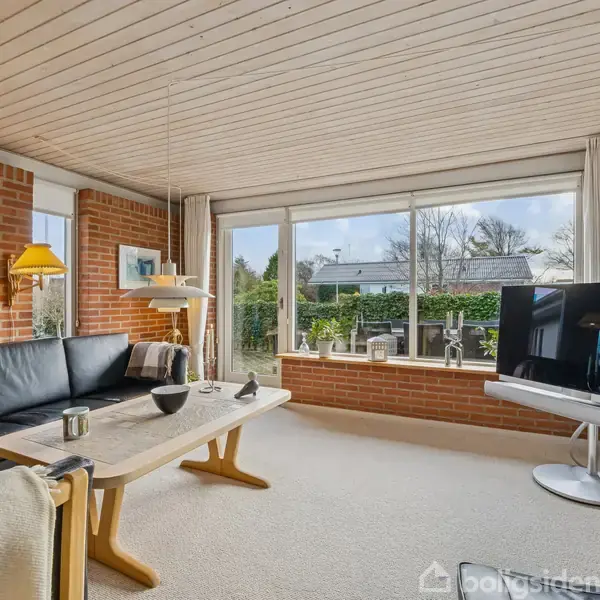 Sort lædersofa står i en stue med murstensvægge, hvor en fladskærms-TV er placeret på en stander. Store vinduer giver udsigt til en velplejet have.