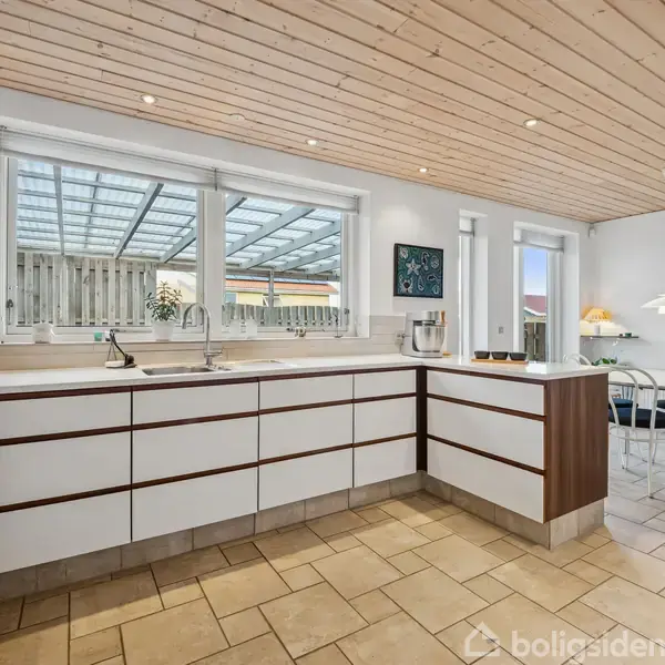 Køkken med hvide skabe og trædetaljer står stille i et rum med lyst klinkegulv og træloft. Store vinduer giver kig til veranda. Rundt bord med stole ses i hjørnet.