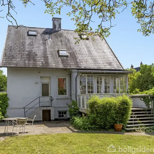 Et hvidt hus med skråt tag står i en grøn have. En trappe fører til en veranda med vinduer. Der er en terrasse med et cafébord og to stole.