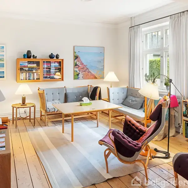 En stue med træmøbler og grå sofa arrangeret rundt om et sofabord. Hylder og billeder hænger på væggen. En stor lampe lyser op i det lyse rum med et trægulv.