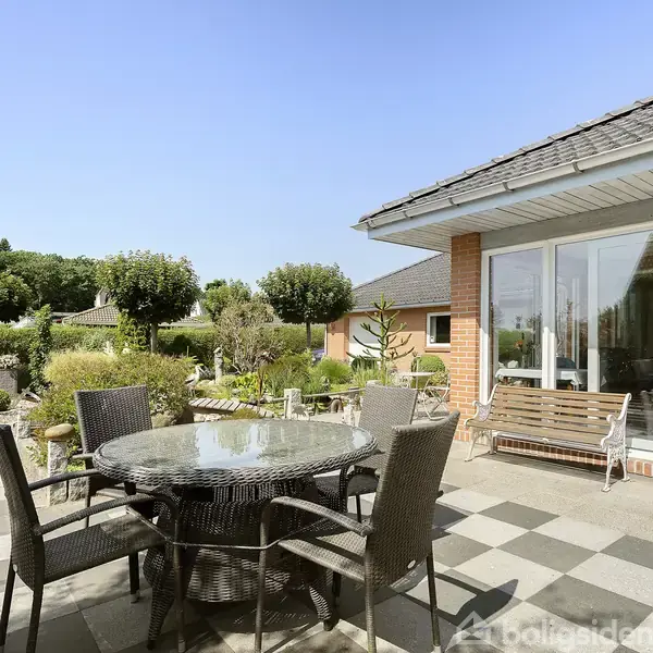 Rundt glasbord med kurvestole står på en flisebelagt terrasse. Ved siden af er et hus med store vinduer, omgivet af en velplejet have med trimmede buske og træer.