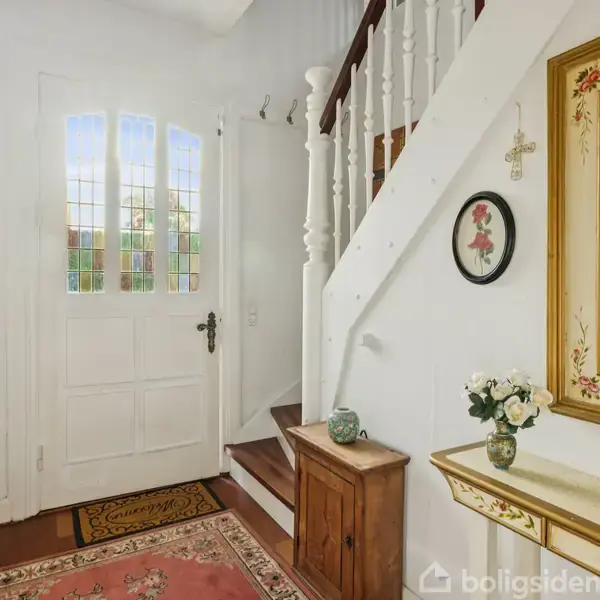 Entrédøren er hvid med glasruder, omgivet af en hvid væg med en trappe til højre. En kommode med blomster og et spejl dekorerer rummet.