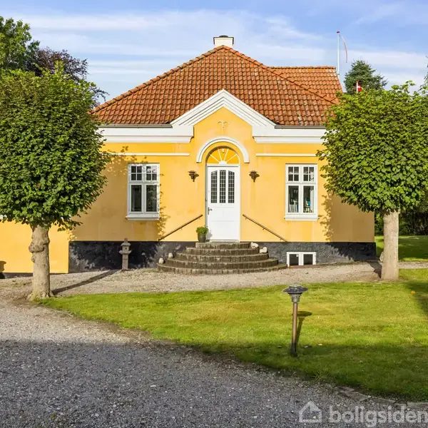 Et gult hus med rødt tag, omgivet af græsplæne og træer, står på en grusvej. Hovedtrappen fører op til en hvid hoveddør i midten.