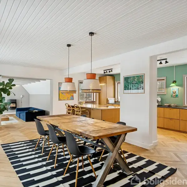 Et rummeligt spisestueområde med et træbord og seks sorte stole står på et sort-hvidt tæppe. Planter, malerier og en åben adgang til køkkenet supplerer rummet.