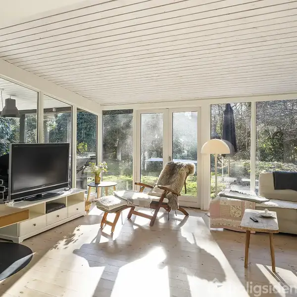 Stue med moderne indretning; stor hjørnesofa og lænestol ved siden af et sofabord; fladskærms-tv foran glasvægge med udsigt til have omgivet af træer.
