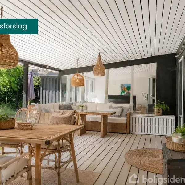 Et lyst, overdækket terrasseområde med træmøbler og kurvelamper. En stor sofa er placeret mod et sortmalet træhus. Tekst: "Indretningsforslag". Grønne planter og tæpper tilfører hygge.