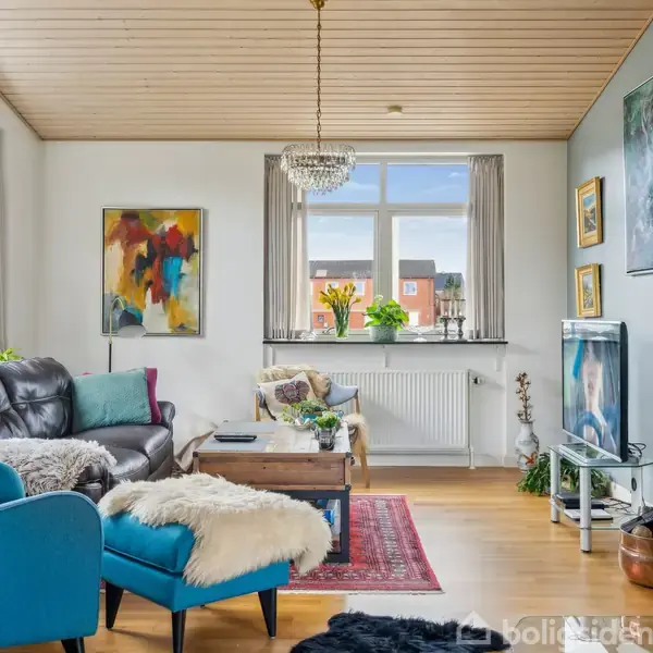 Stue med blå og sort sofa, sofabord, tæppe og kunst på væggen. Brændeovn til højre, billeder og planter i vinduet. Lyse lofter og træplankegulv skaber hyggelig atmosfære.