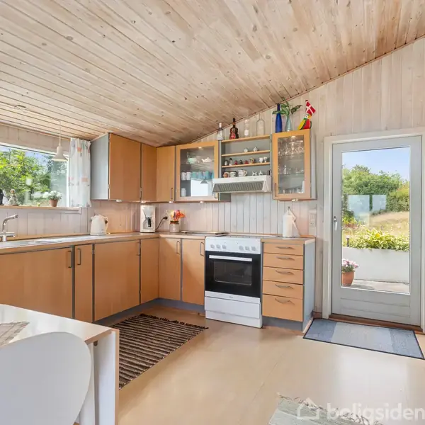 Køkken med træloft og træskabe, hvor en ovn og vask er i brug. Indretningsartikler placeres på hylder, og en dør fører ud til en have belyst af naturligt lys.