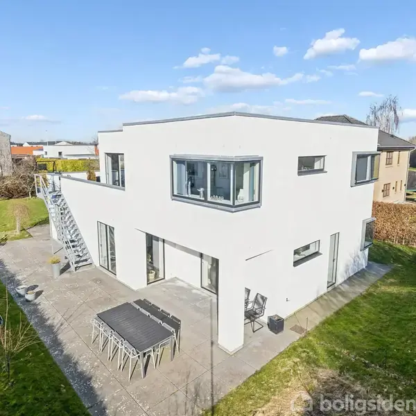 Moderne, hvidt hus med fladt tag har store vinduespartier og udendørs spiseområde på stenterrasse. En udvendig trappe fører til øverste etage. Haven er omgivet af hække.