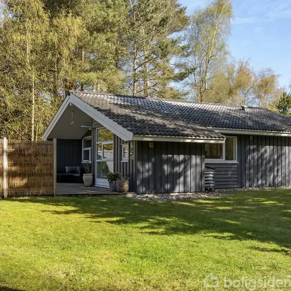 Et træsommerhus står stille i en grøn have omgivet af skov. Huset har gråt træværk og tag, med glasdør og vinduer mod venstre. Ingen tekst synlig.