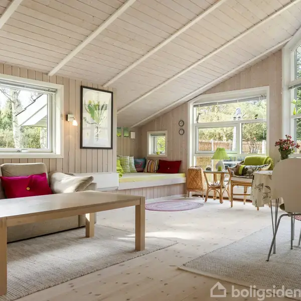 Stue med skråt loft, hvor lyse trævægge og store vinduer skaber en lys atmosfære. Sofa med farverige puder, træbord og spiseområde med stole. Grøn udsigt udenfor.