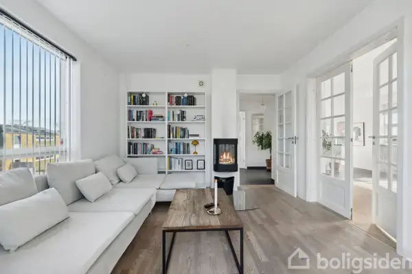 En hvid sofa står ved siden af en bogreol med bøger og dekorationer; et tændt ildsted står på bagvæggen. Rummet er lyst med trægulve og glasdøre.