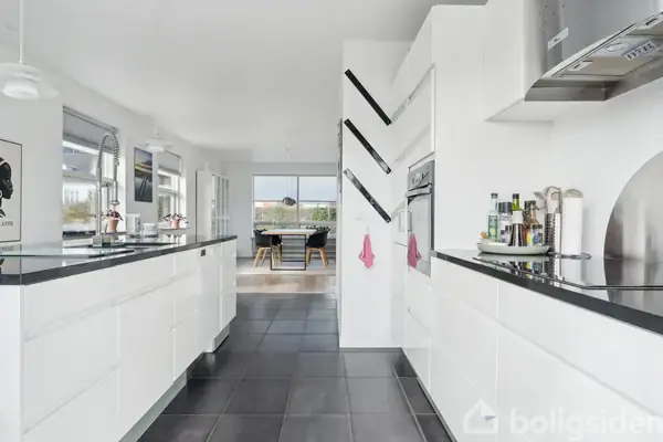 Et moderne køkken med hvide skabe og sorte bordplader. Rummet er lyst med store vinduer og sort gulvflisebelægning. I baggrunden ses en åben spisestue med et stort vindue.