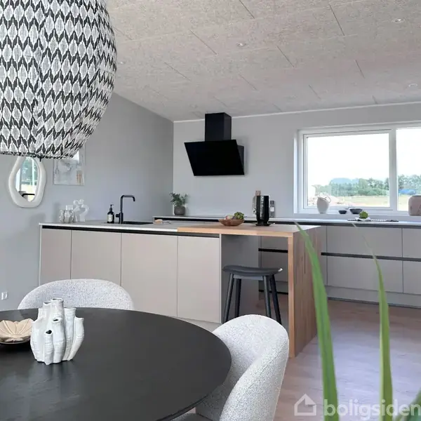 Rundt spisebord med to hvide stole i forgrunden, en dekorativ lampe hænger over det. Moderne køkken bagved med lyse skabe, sort emhætte og stort vindue med udsigt.