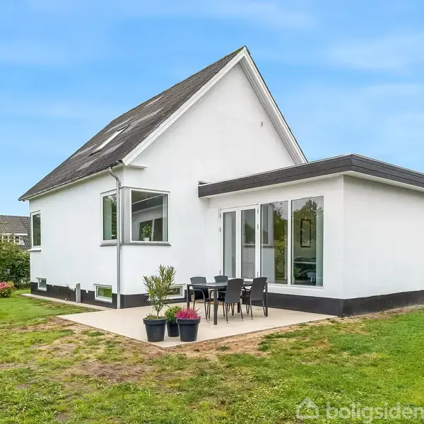 Hvidt hus står stille på græsplæne med terrasse i forgrunden, hvor sorte havemøbler står under åben, blå himmel. Ingen tekst til stede.