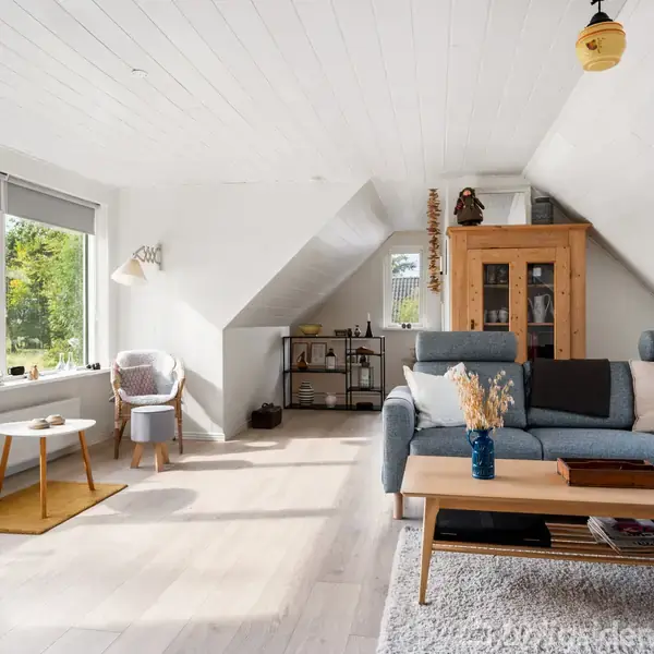 Stue med blå hjørnesofa med puder, sofabord, og skænk i et lyst rum. Store vinduer og skråvægge giver udsigt til grøn natur udenfor. Hylder og planter dekorerer rummet.