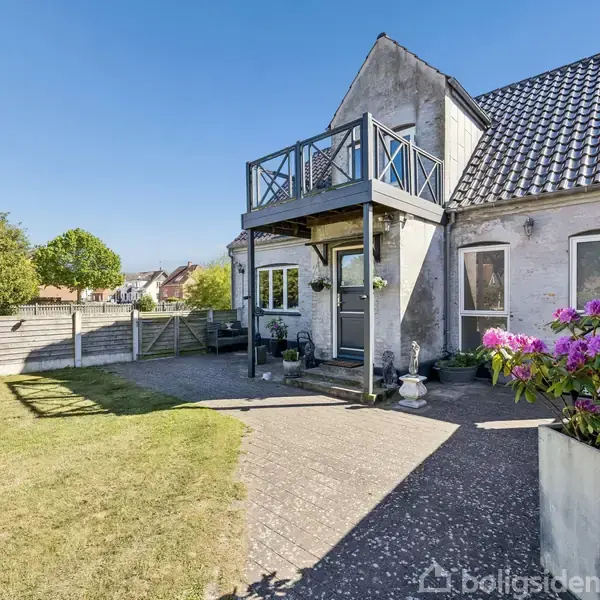 Huset står i en gårdhave med blomster i en potte og en flisebelagt sti foran. Over indgangen er der en balkon. Omkring er der en grøn græsplæne og et hegn.