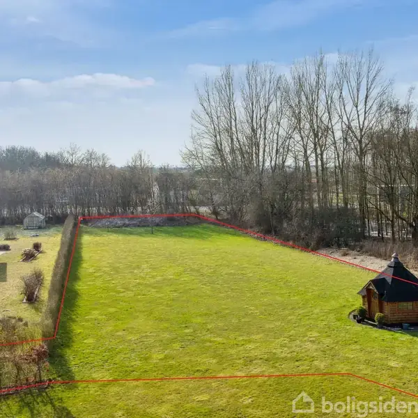 Græsplæne med en lille, sekskantet træpavillon; omgivet af høje træer og hegn, med et nærliggende hus til venstre. Sollys oplyser området under en delvist skyet himmel.