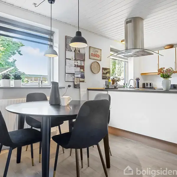 Et moderne køkken med en rund sort spisebord omgivet af fire stole. Baggrunden består af hvidt interiør, en emhætte over kogepladen, planter på køkkenbordet og en vægplakat med teksten "Sorry, we're closed."