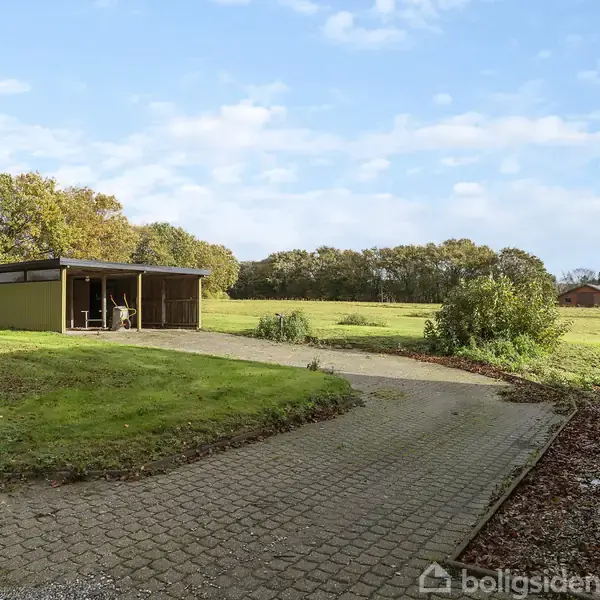 Et lille udhus står på en græsplæne, flankeret af en flisebelagt sti. Omgivelserne består af en stor, åben mark med træer i baggrunden under en klar blå himmel.