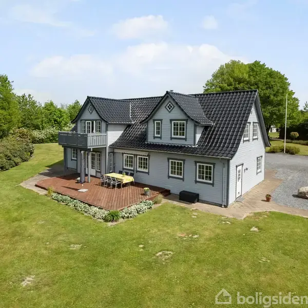 To-etagers hus med sort tag og hvide vægge står omgivet af en grøn græsplæne og træer. En træterrasse med havemøbler er foran huset.
